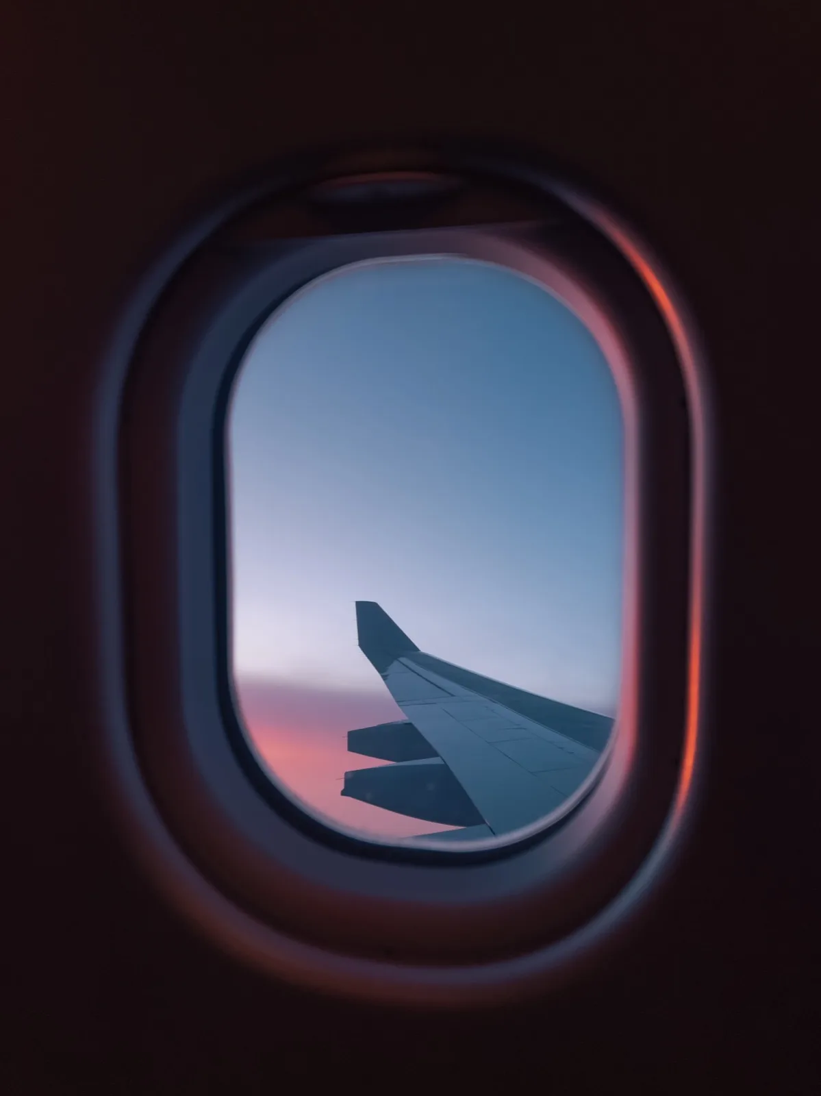 An airplane wing seen through a passenger window against a pale blue sky with a pink horizon