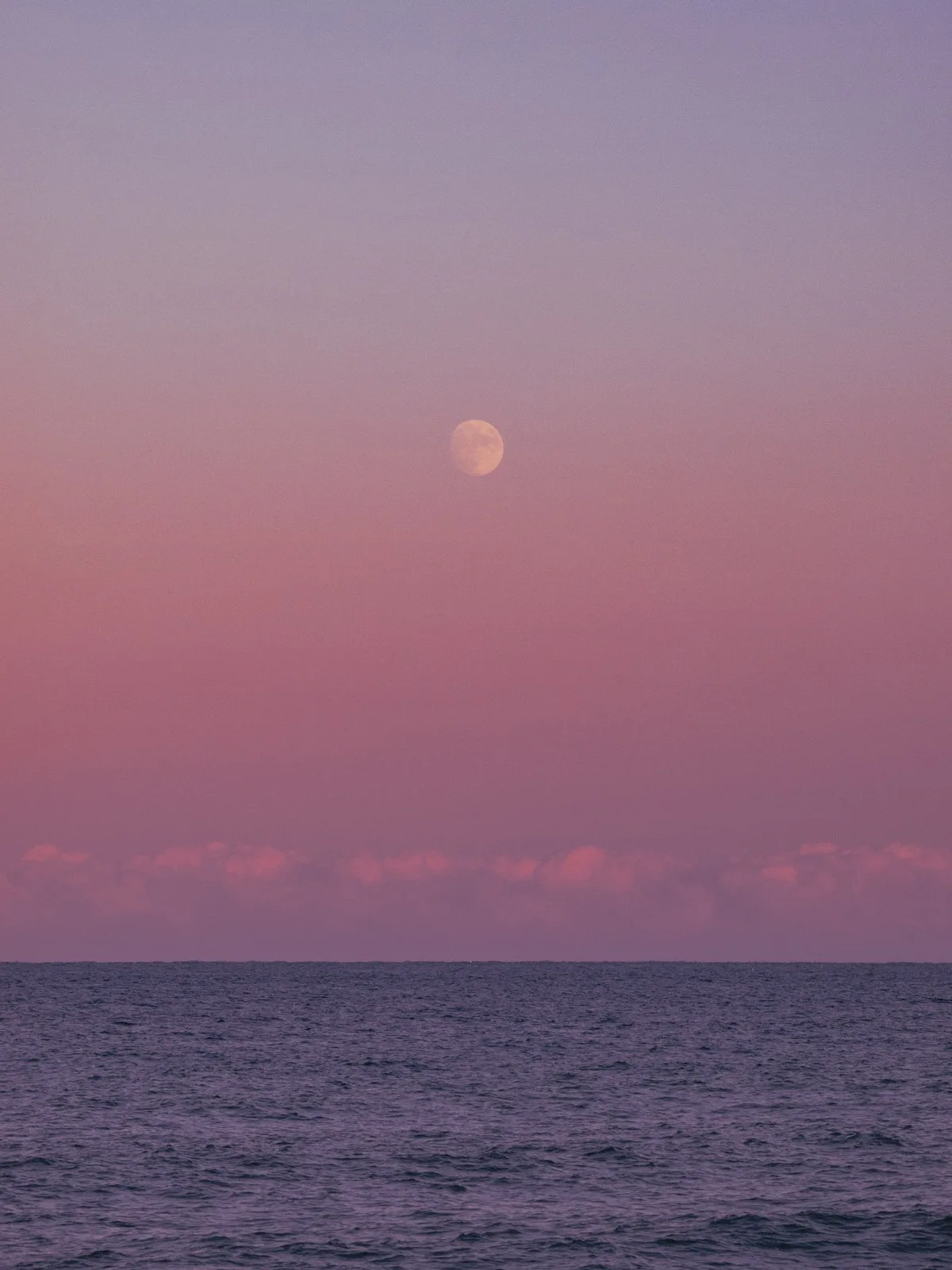 The moon hanging above a calm dark sea under a soft pink and lavender dusk sky