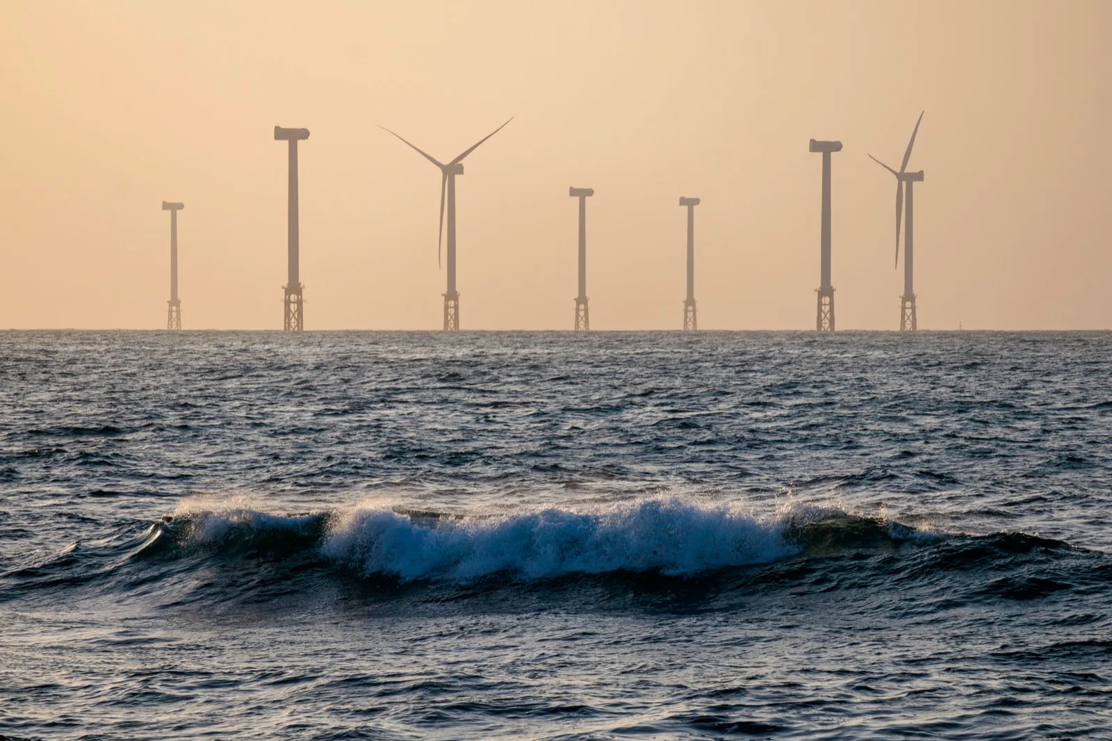 Offshore wind turbines lined up beyond dark ocean waves in hazy evening light