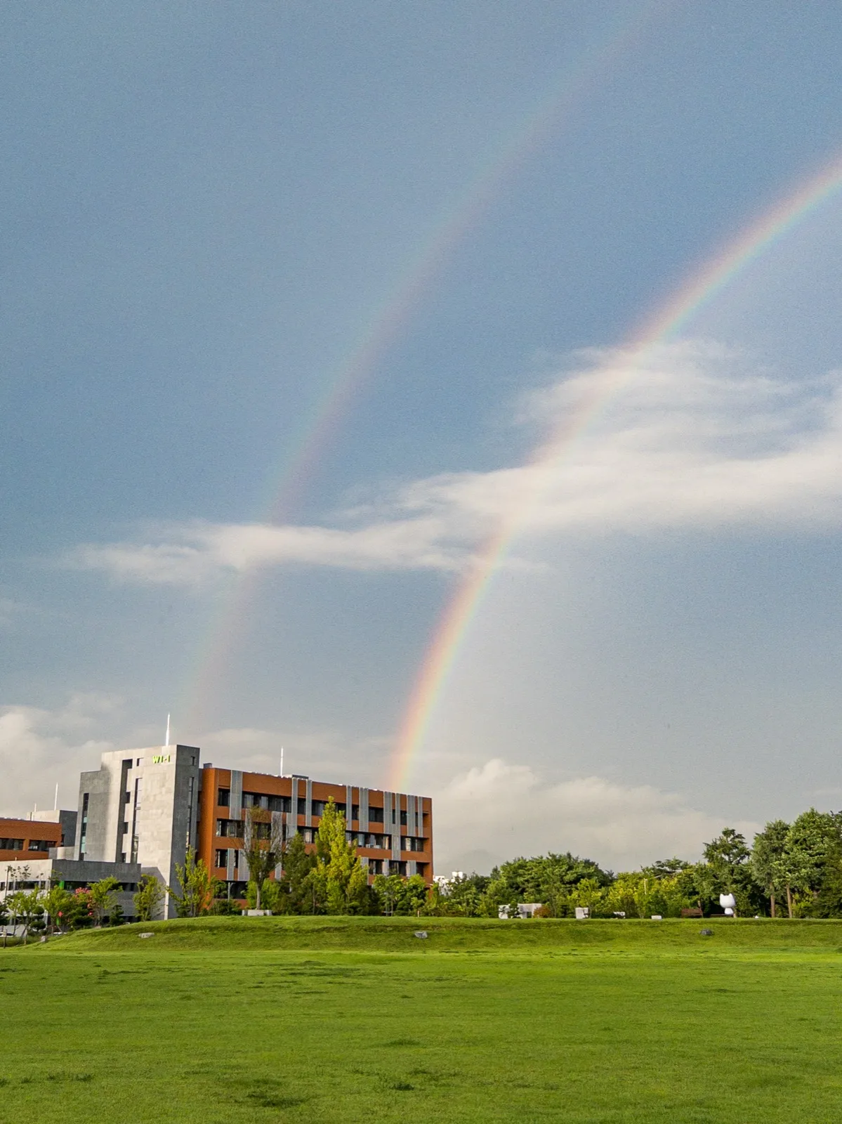 A double rainbow arching above a grassy field and campus buildings after rain