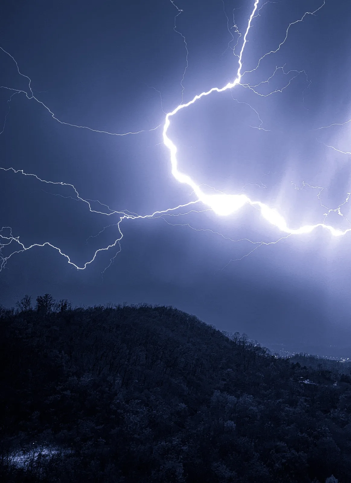 A bright forked lightning strike spreading across the night sky above a dark wooded ridge