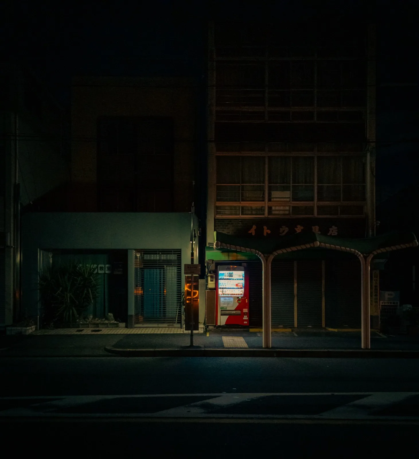 A brightly lit vending machine glowing on a dark, quiet street lined with shuttered buildings at night