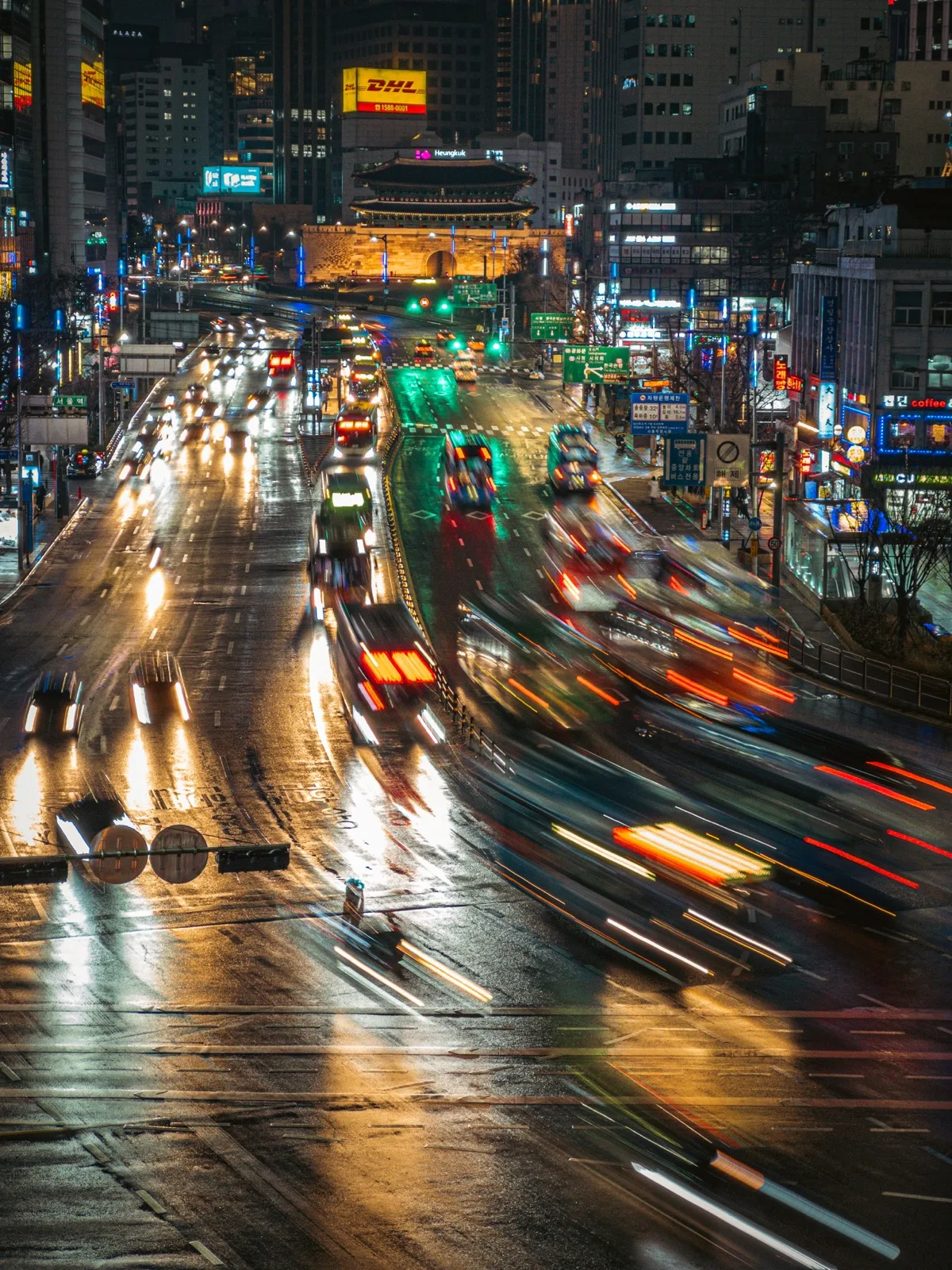 Night traffic streaking along a wet city avenue toward Sungnyemun Gate glowing in the distance