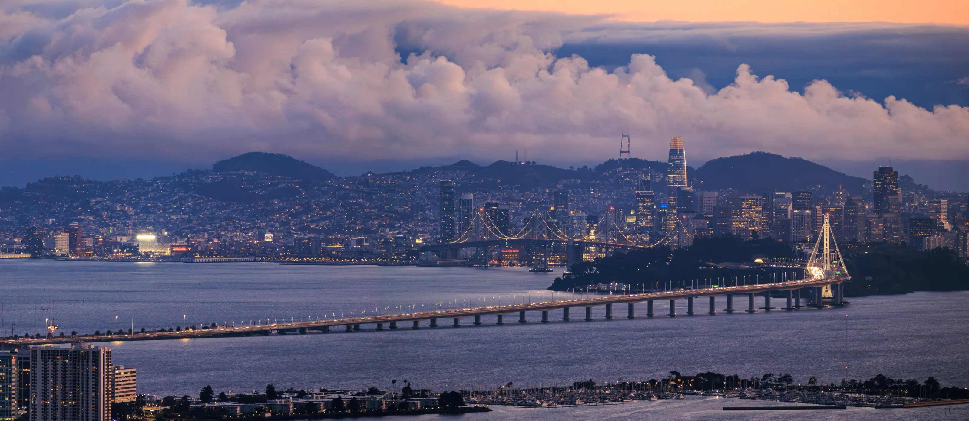 A panoramic dusk view of San Francisco and the Bay Bridge under pink clouds, with city lights along the bay