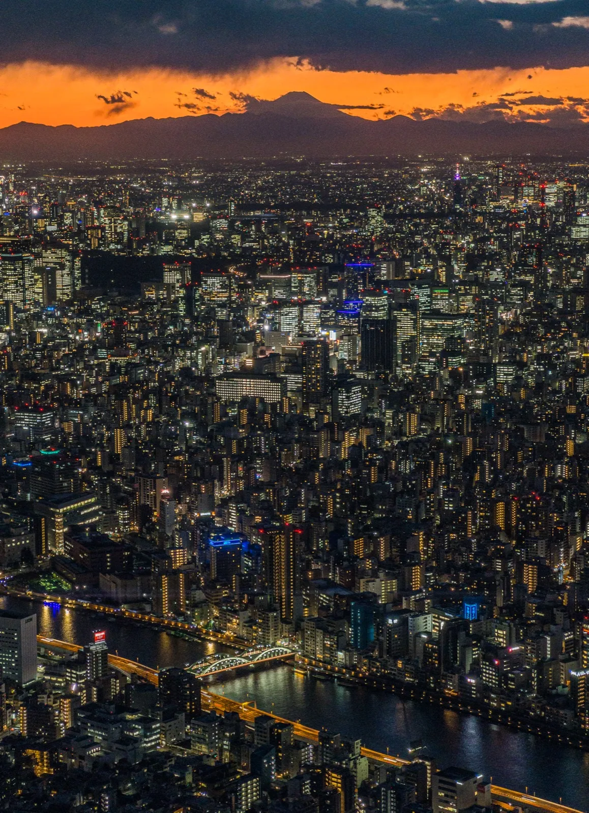 A dense Tokyo cityscape at dusk with a river in the foreground and mountains silhouetted under an orange sky